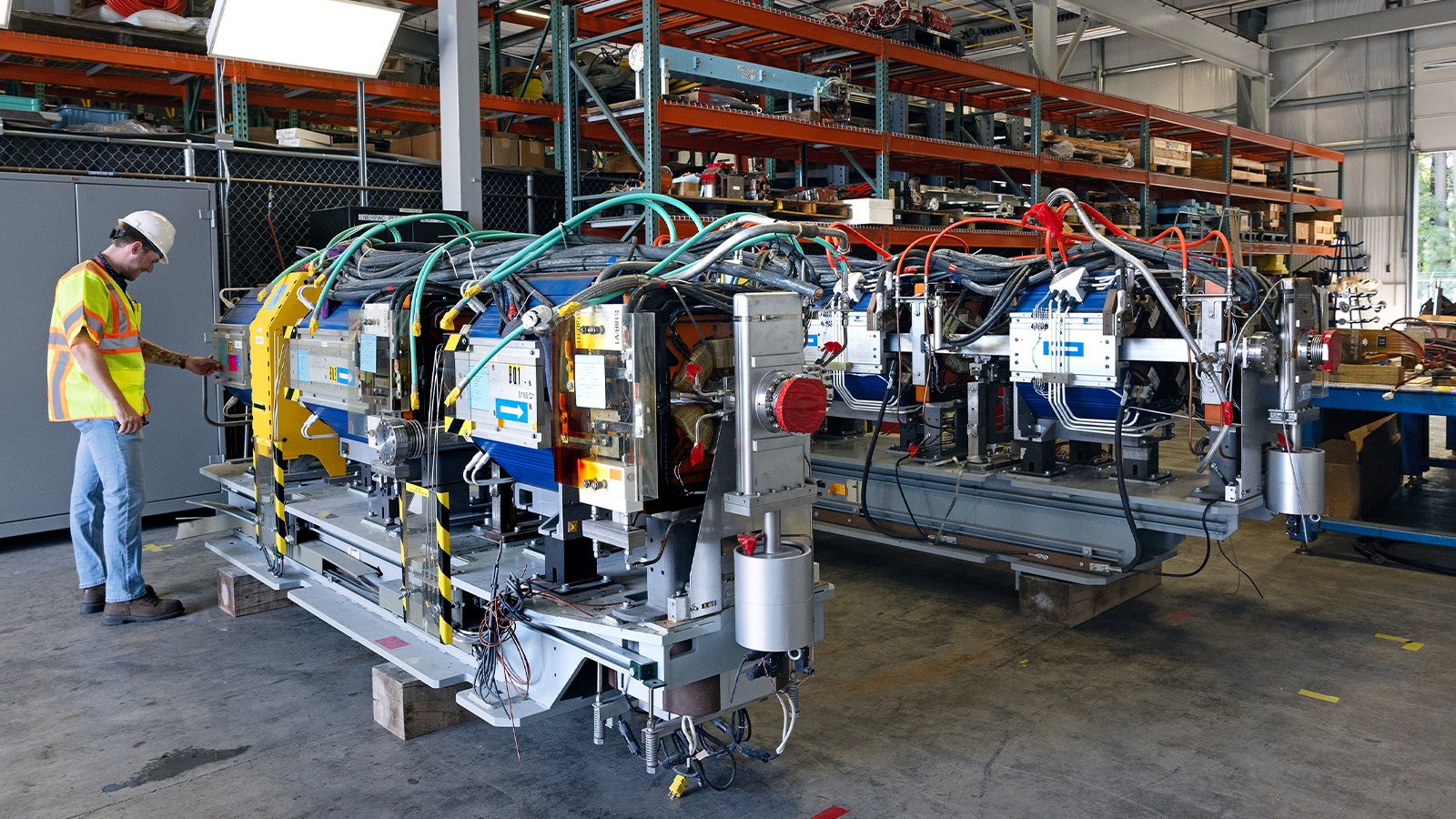 Two modules of brightly colored magnets and wires sit next to each other in a storage unit at Thomas Jefferson National Laboratory as a worker in a safety vest examines one of them.