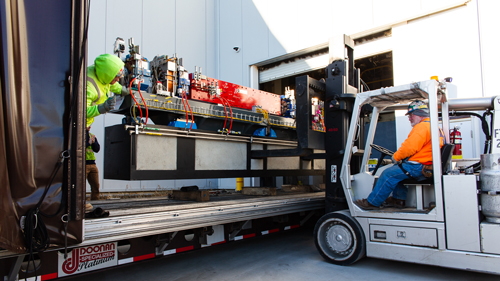 A worker in a forklift truck helps another to move a heavy module of magnets through a door. 